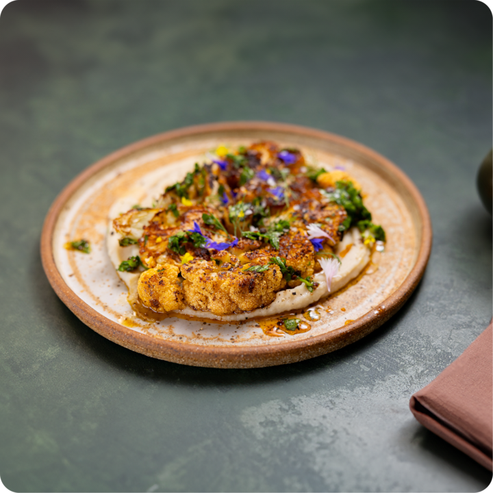 Un steak de chou-fleur rôti garni d'herbes fraîches, de fleurs comestibles colorées et d'épices, servi sur un lit de sauce blanche crémeuse, présenté dans une assiette en céramique sur une surface verte texturée.