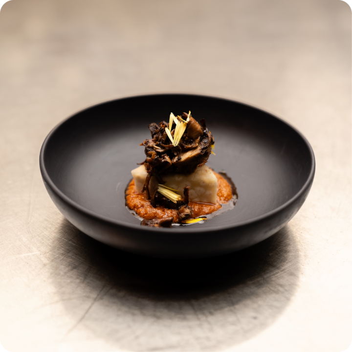 Un plat gastronomique composé d'une portion carrée de polenta blanche surmontée de champignons sautés, garnie de pétales jaunes, servie sur un lit de sauce rouge dans un bol noir.