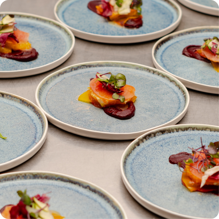 Plan rapproché de plusieurs assiettes bleues contenant des plats gastronomiques composés de fines tranches de saumon, de purée de betterave, de micropousses et de garnitures colorées.