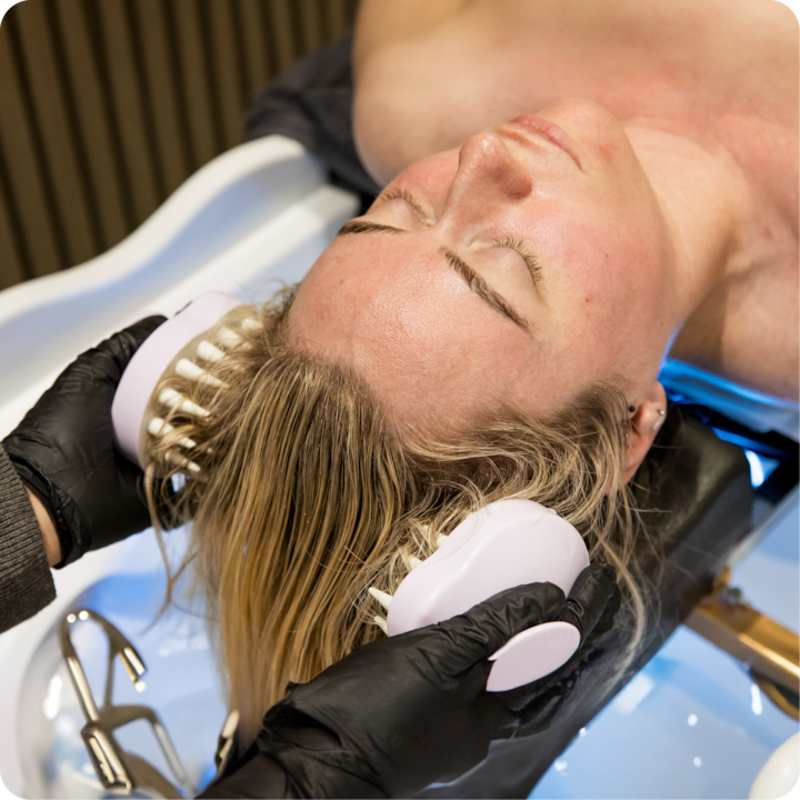 Plan rapproché d'une coiffeuse portant des gants noirs et utilisant un outil de massage du cuir chevelu blanc sur les cheveux blonds mouillés d'une cliente au-dessus d'un lavabo de salon.