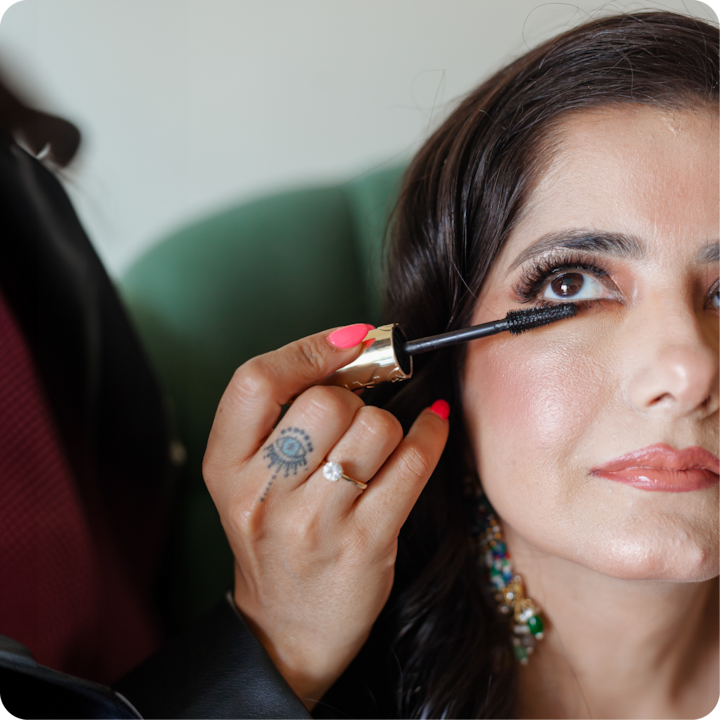 Une main portant un tatouage d'œil et une bague en diamant applique du mascara sur les cils inférieurs d'une personne, avec une chaise verte en arrière-plan.