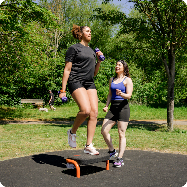 Deux femmes font de l'exercice dans un parc. L'une d'elles monte sur une plateforme orange et noire et tient des haltères. Elles sont entourées d'une végétation luxuriante et d'arbres.