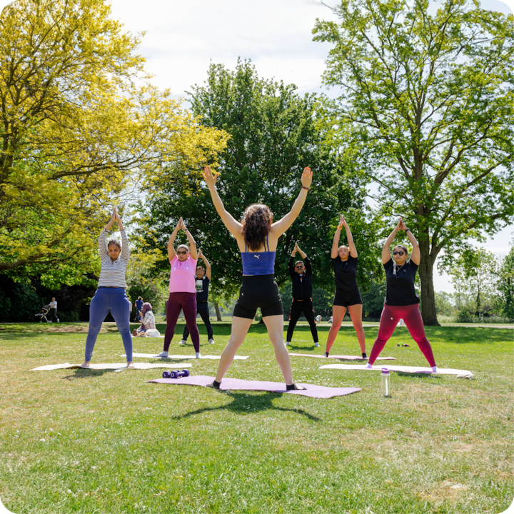 Un groupe de personnes participent à une séance de yoga en plein air dans un parc ensoleillé, avec des arbres et de l'herbe en arrière-plan. Un professeur de yoga se tient à l'avant, guidant les participants qui s'étirent en levant les bras au-dessus de la tête.