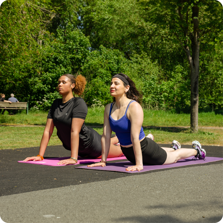 Deux personnes pratiquent le yoga dans un parc en plein air. Elles exécutent la posture du chien tête en haut sur des tapis de yoga roses, entourées d'arbres et d'herbe par une journée ensoleillée.