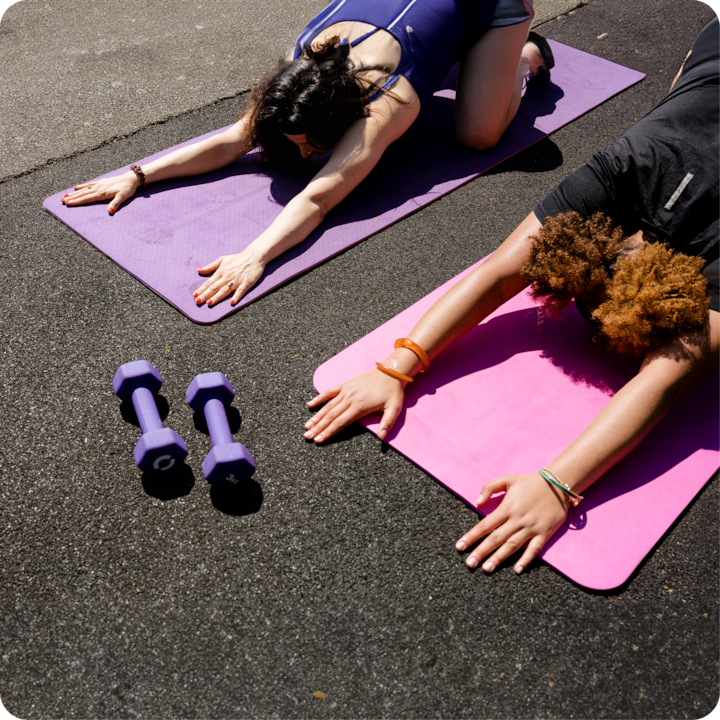 Deux personnes font du yoga sur des tapis violets et roses à l'extérieur par une journée ensoleillée, avec une paire d'haltères violets posés sur l'asphalte à proximité.