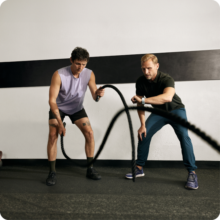 Deux personnes participent à une séance d'entraînement avec des cordes ondulatoires dans une salle de sport, l'une porte un short de sport et un débardeur et l'autre un polo et un pantalon de sport, contre un mur uni avec une bande noire.