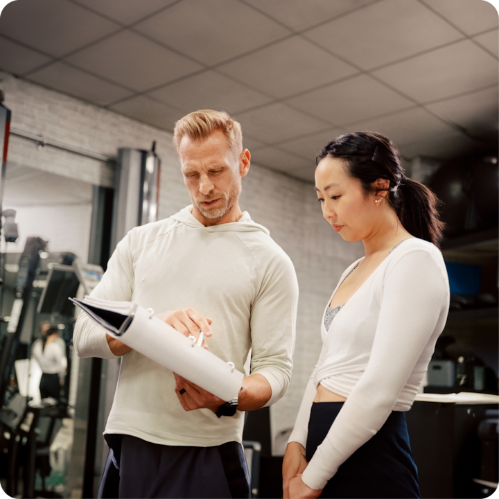 Un entraîneur de fitness tenant un porte-bloc discute d'un programme d'entraînement avec une personne dans une salle de sport. Ils sont entourés d'équipements d'exercice et d'un mur en miroir.
