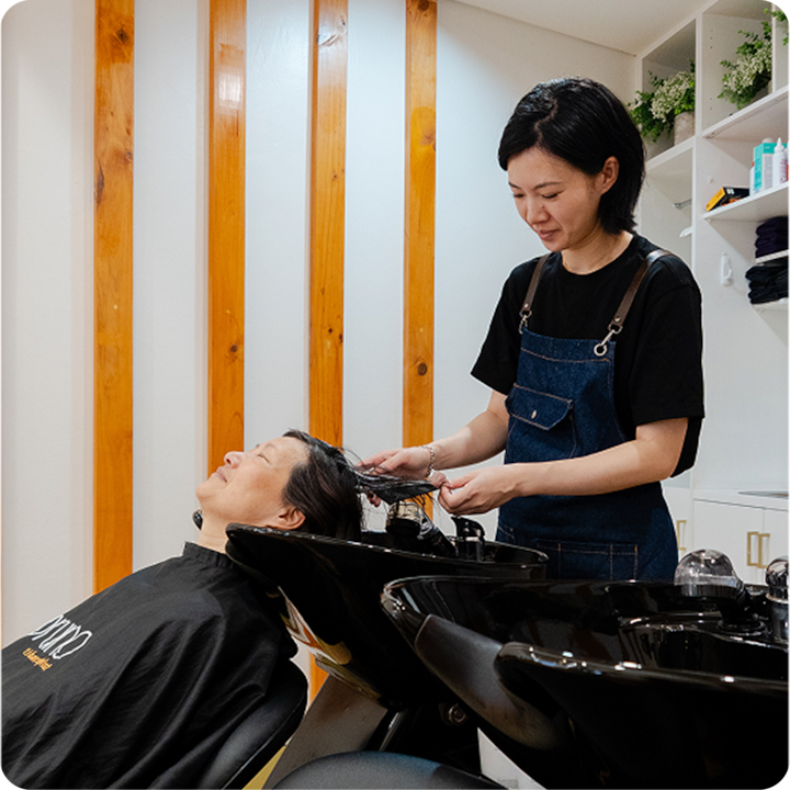 Une coiffeuse portant un tablier en jean lave les cheveux d'une cliente au-dessus d'un bac de salon de coiffure noir, dans une pièce moderne et bien éclairée avec des accents en bois au mur.