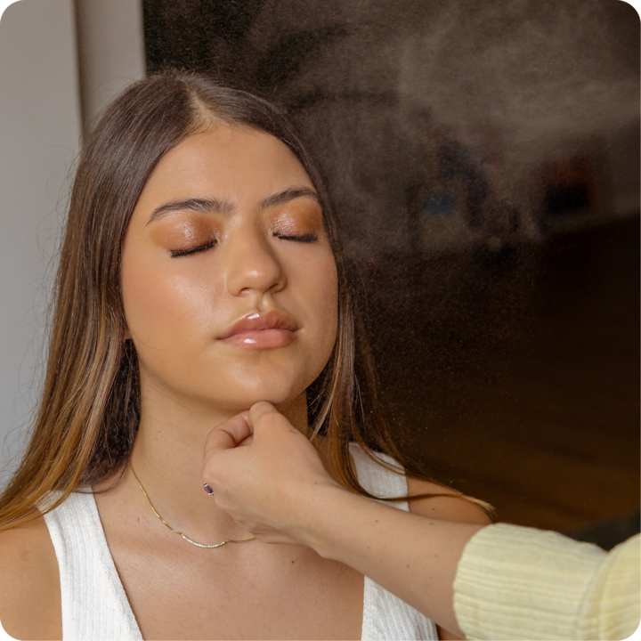 Image en gros plan montrant une main qui applique un spray de beauté près du cou et de la clavicule d'une personne portant un débardeur blanc et un collier doré.