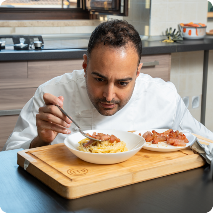 Une planche à découper en bois sur une table avec un plat de pâtes garni de morceaux de bacon croustillants, à côté d'une assiette remplie de tranches de bacon en plus. Un plan de travail et une cuisinière sont visibles en arrière-plan.