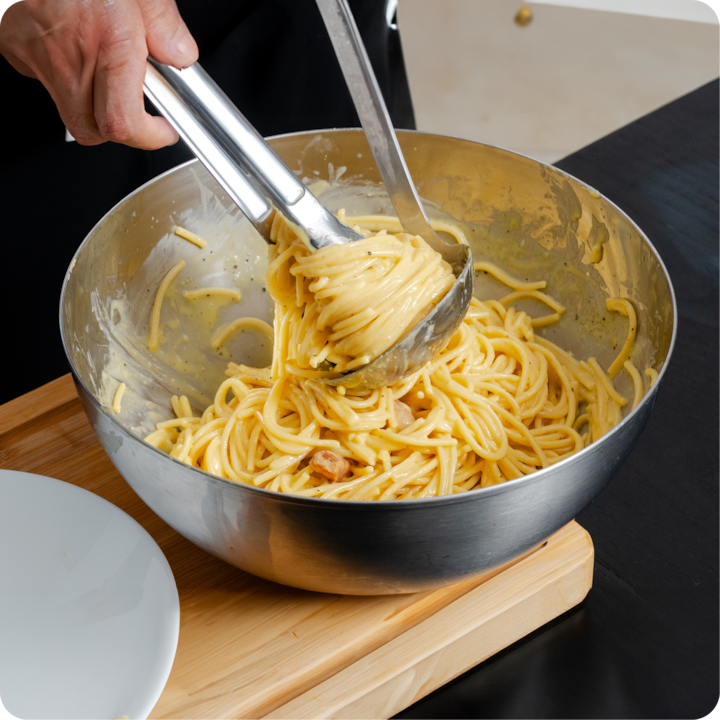 Plan rapproché d'une personne utilisant des pinces et une louche pour prendre des spaghettis à la crème dans un saladier en métal, mettant en évidence les pâtes fraîchement préparées.