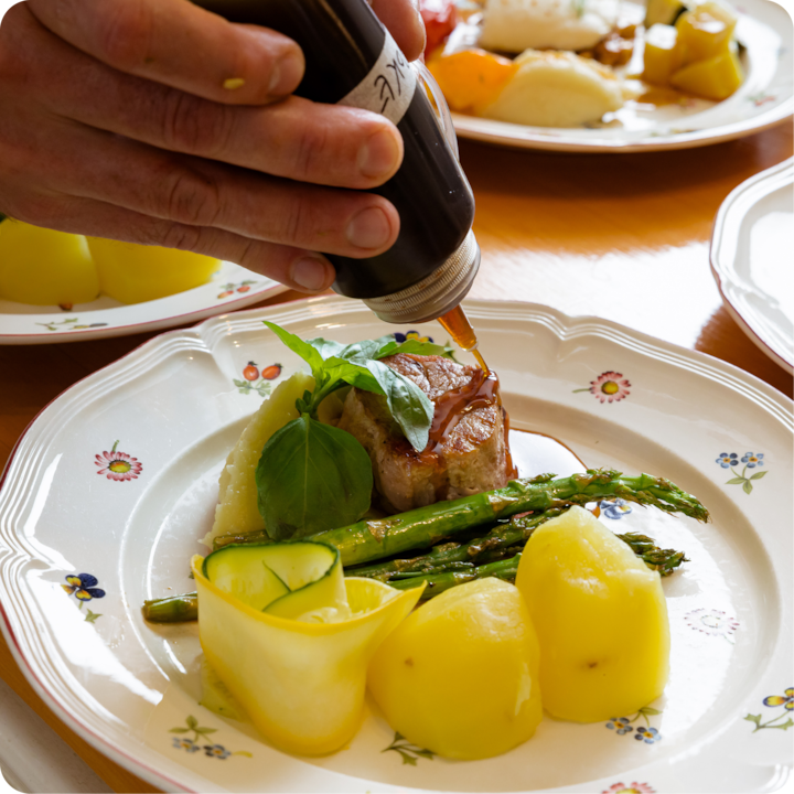 Plan rapproché d'un plat composé d'asperges grillées, de pommes de terre dorées, de lanières de courgettes et d'un morceau de viande poêlée garni d'une feuille de basilic, tandis qu'une main verse de la sauce sur la viande.