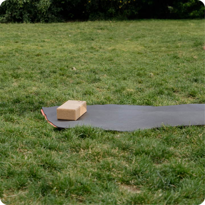 Bloc de yoga en liège posé sur un tapis de yoga noir étendu sur une pelouse, évoquant un environnement d'entraînement calme en plein air.