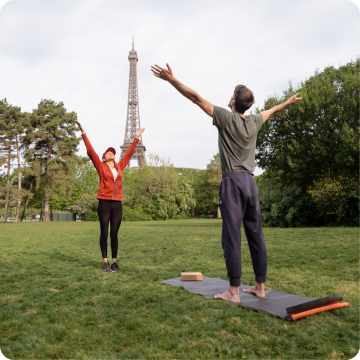 Deux personnes entourées d'arbres font des étirements de yoga sur une étendue d'herbe près de la tour Eiffel avec des tapis de yoga et un bloc de mousse.