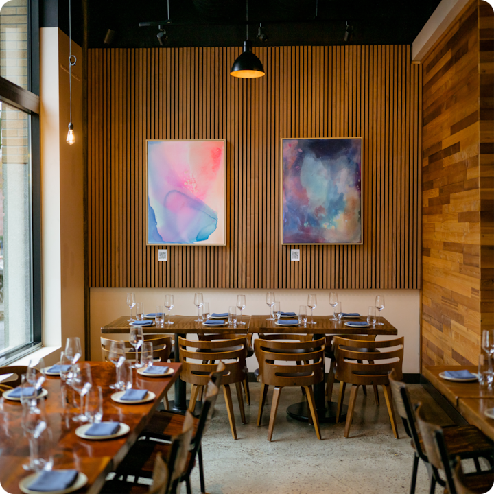 Coin repas moderne figurant une table en bois dressée avec des verres et des assiettes soigneusement disposés. Deux peintures abstraites colorées sont accrochées à un mur à lattes de bois en arrière-plan.