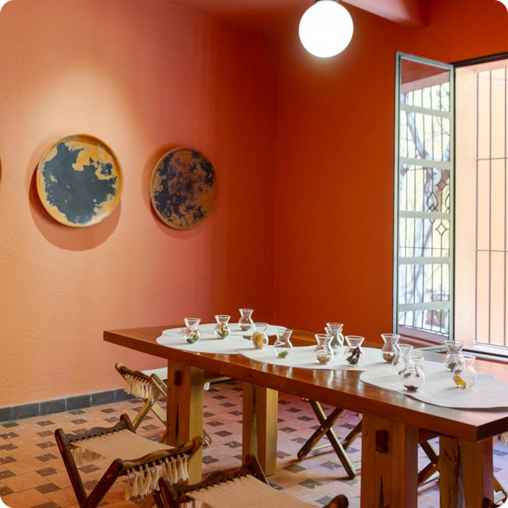 Une pièce chaleureuse aux murs orange avec une longue table en bois dressée pour une dégustation, avec de petits bols en verre remplis de divers ingrédients et des chaises rustiques en tissu placées autour de la table ; trois plats décoratifs sont accrochés au mur et les rayons du soleil entrent par une fenêtre ouverte.