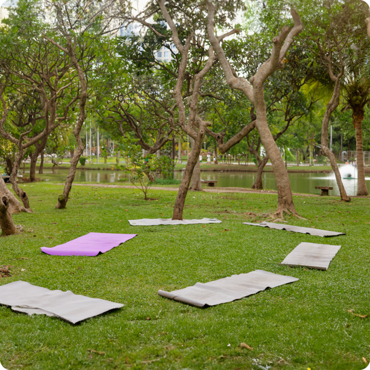 Un cadre extérieur paisible avec des tapis de yoga disposés en cercle sur de l'herbe verte, des arbres et un étang en arrière-plan.