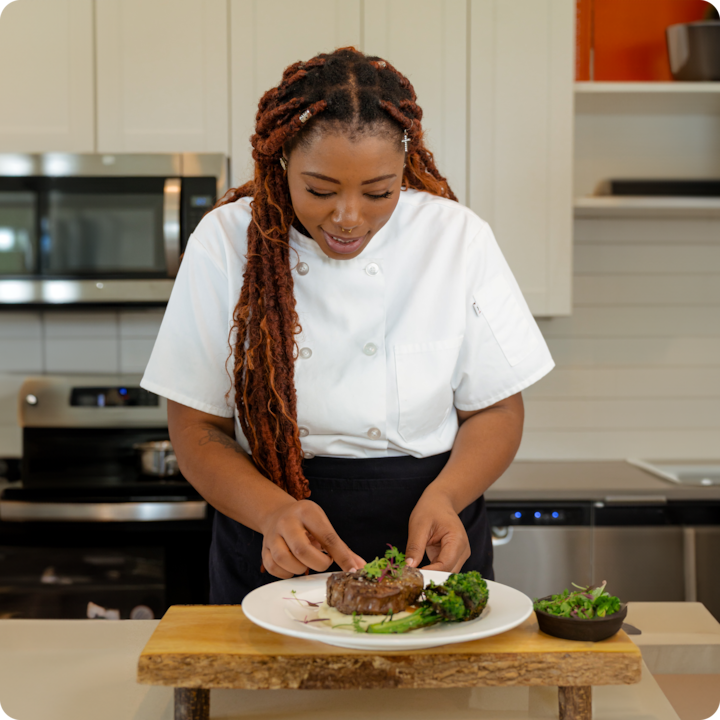 Dans une cuisine moderne, une cheffe vêtue d'une tenue blanche dresse un steak accompagné de légumes rôtis et de pousses sur une planche en bois.