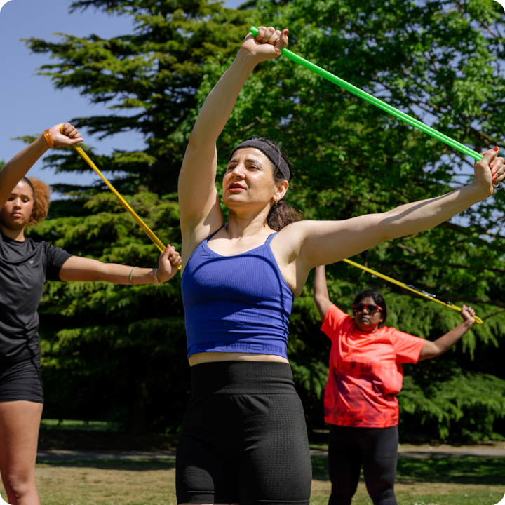 Un groupe de personnes participent à un cours de fitness en plein air dans un parc. Elles utilisent des bandes de résistance colorées sous un ciel bleu clair avec des arbres en arrière-plan.