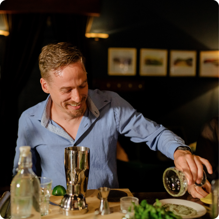 Une personne vêtue d'une chemise bleue se tient derrière un bar, versant le liquide d'une petite carafe en verre dans un shaker. Le comptoir du bar est garni d'ingrédients, notamment un citron vert, une bouteille de liquide transparent et des herbes fraîches. L'arrière-plan présente des murs sombres et des œuvres d'art encadrées, créant une atmosphère chaleureuse.