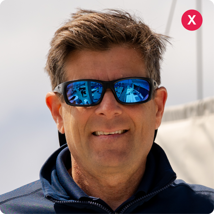 Gros plan d'une personne portant des lunettes de soleil et une veste zippée sombre, debout en extérieur avec une voile floue et un ciel nuageux en arrière-plan.