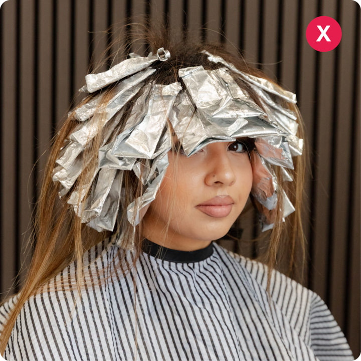 Une personne est assise dans le fauteuil d'un salon de coiffure devant un arrière-plan en lambris. Elle porte une cape rayée, ses cheveux sont enveloppés dans des feuilles d'aluminium pendant une coloration.