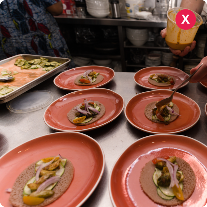 Plusieurs assiettes rouges sur un comptoir, chacune contenant un pain plat injera garni de légumes frais comme des courgettes, des tomates et des oignons rouges, tandis qu'une personne verse de la sauce sur une assiette dans une cuisine professionnelle.