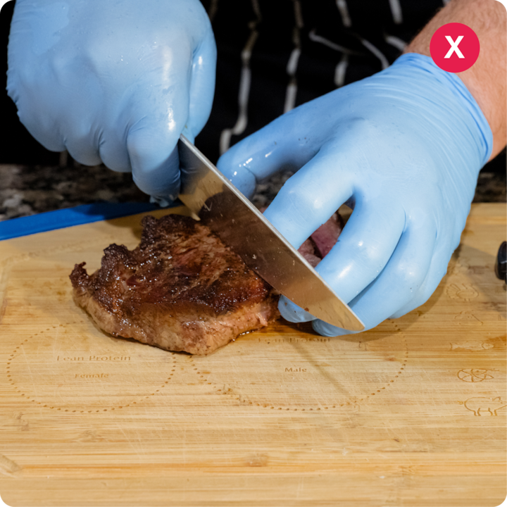 Plan rapproché sur des mains portant des gants bleus qui tranchent un steak cuit sur une planche à découper en bois présentant des incisions.