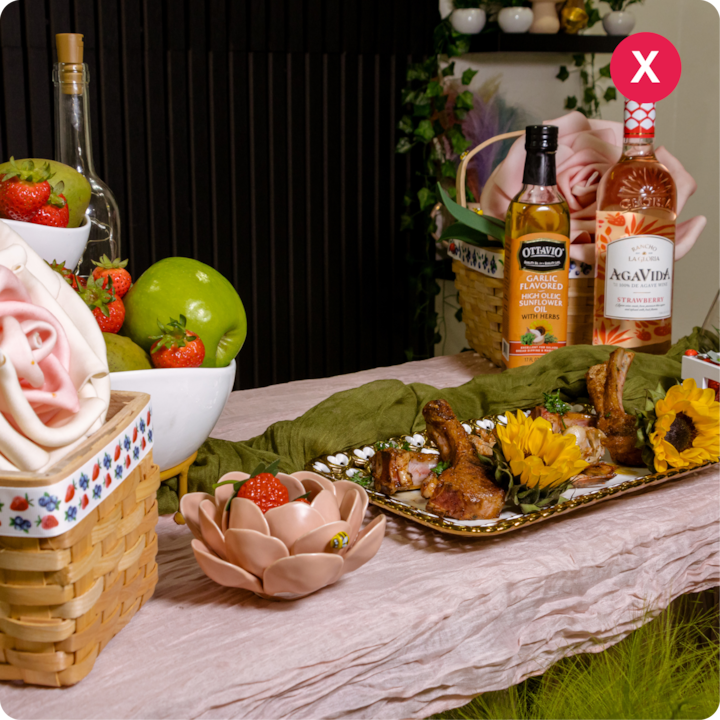 Une table décorée avec un plateau de côtelettes d'agneau grillées garnies de tournesols, entouré de fraises fraîches, de pommes vertes, d'une bouteille d'huile d'olive aromatisée à l'ail et d'une bouteille de liqueur de fraise sur une nappe rose avec un chemin de table vert.