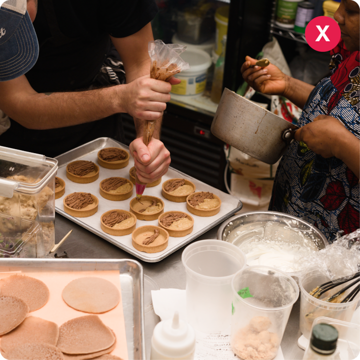 Deux personnes travaillent ensemble dans une cuisine. L'une dresse des fonds de tartelettes sur un plateau avec une poche à douille remplie de chocolat, tandis que l'autre tient une casserole. L'espace de travail est couvert d'ustensiles de pâtisserie et d'ingrédients, notamment de la pâte et de la crème fouettée.