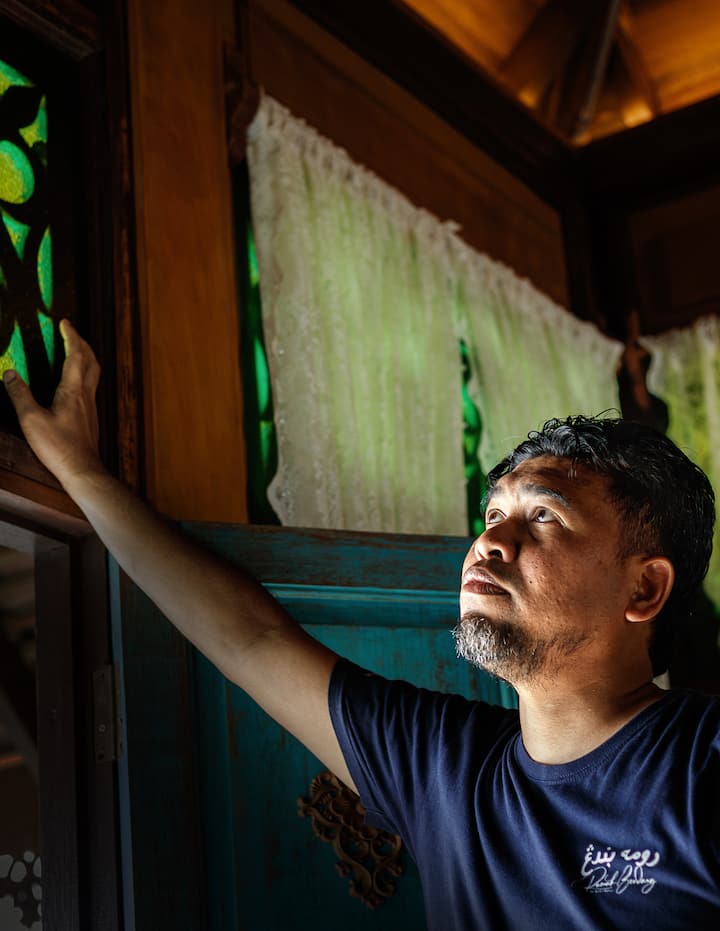 A person stands in a traditional Malaysian wooden home, with intricate green stained-glass windows and lace curtains in the background, showcasing cultural heritage.