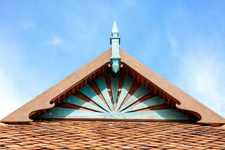 A traditional Malay gable roof made of reclaimed timber, featuring a blue pointed ornament and sunlight pattern, set against a clear blue sky.