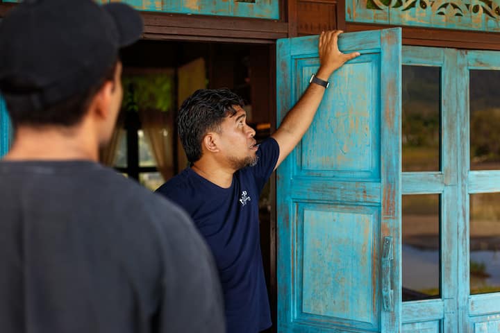 A man stands holding the edge of a vibrant turquoise wooden door, gesturing towards the open space of a traditional-style home with a scenic outdoor view in Langkawi.