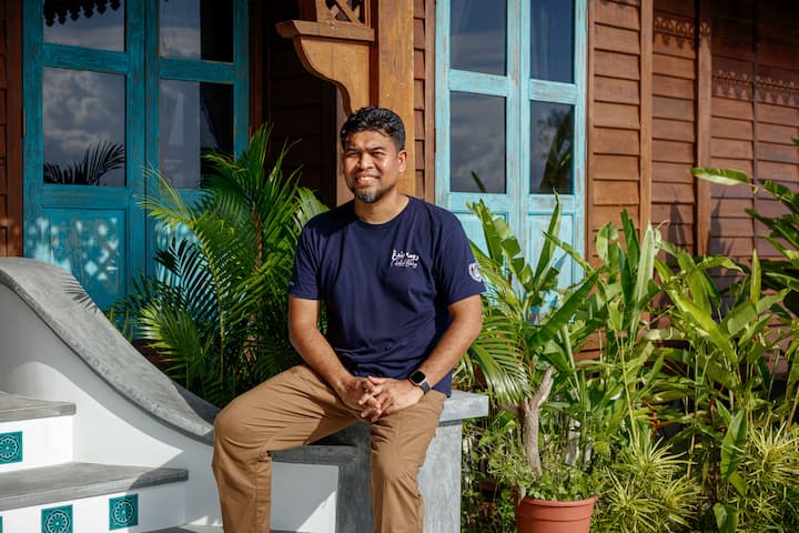 A person seated on a stone bench in front of a traditional wooden house with turquoise window frames, surrounded by lush greenery including potted plants and palm leaves.