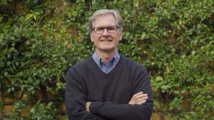 A person standing with arms crossed in front of a lush green ivy-covered wall, wearing a black sweater over a blue collared shirt.