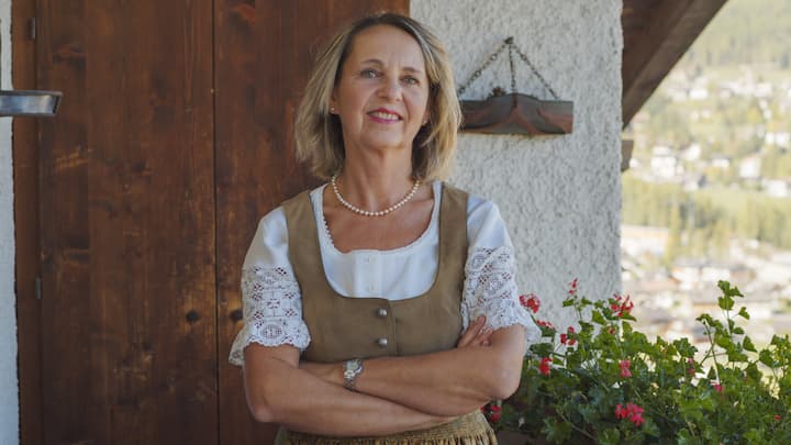 A host wearing traditional Alpine attire stands with arms crossed in front of a rustic wooden door, with red flowers and a scenic mountainous landscape visible in the background.