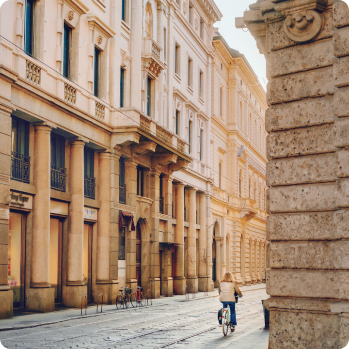 A quiet, marble clad street corner in Milan. A woman wearing a warm winter coat bikes into the distance.