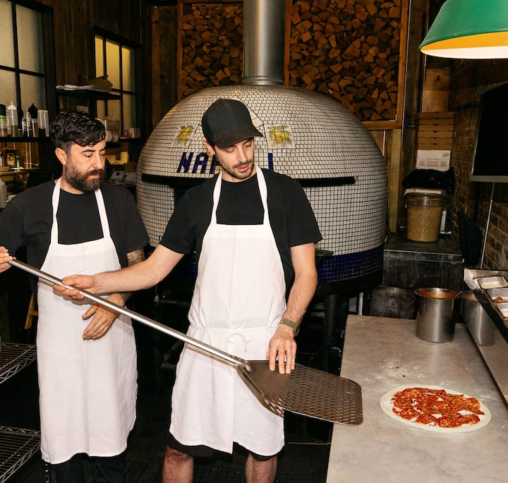 An image of two people making pizza next to a wood-fired oven