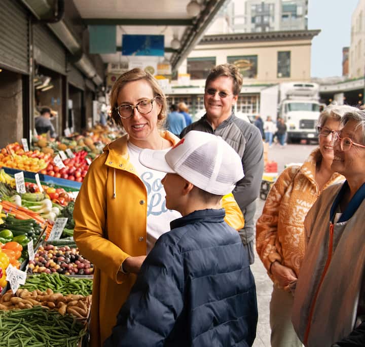 Պատկերում՝ մի խումբ մարդիկ, որոնք հանրային շուկայում վայելում են վաճառասեղանի վրայի մթերքը