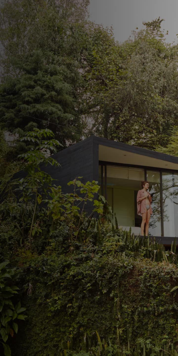 Žena stoji na terasi moderne kuće izgrađene na padini. U rukama naslonjenim uz prsa drži šalicu kave i gleda u daljinu. Padina je prekrivena bujnim zelenilom koje se obavija oko kuće.