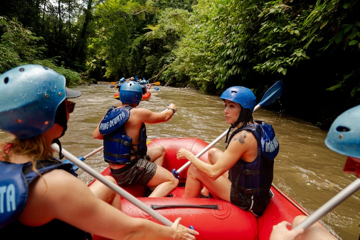 Un groupe de personnes portant des casques bleus et des gilets de sauvetage pagayant sur un canoë gonflable rouge le long d'une rivière étroite entourée d'une jungle verdoyante.