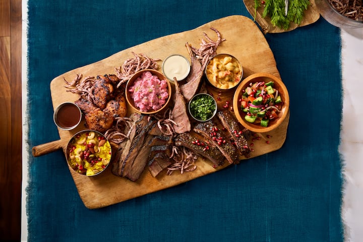 Une planche en bois avec un assortiment de viandes grillées, des légumes colorés et diverses sauces sur une nappe bleu foncé.