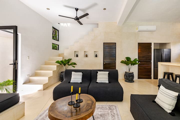 Salon moderne aux murs beiges avec un canapé noir décoré de coussins rayés, une table basse en bois avec trois bougeoirs et un escalier ouvert menant à l'étage. De grandes plantes en pot et la lumière naturelle mettent en valeur cet espace.