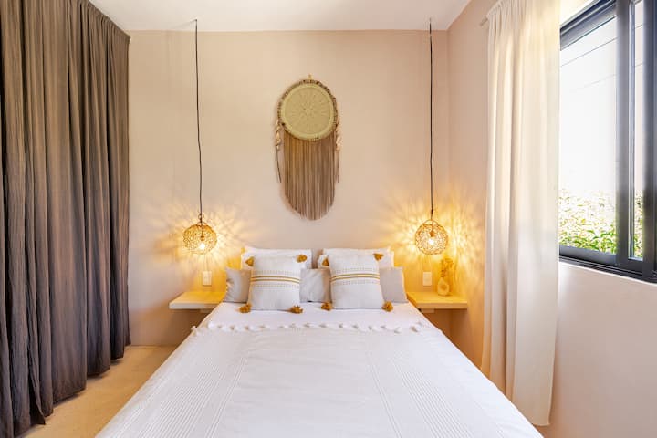 Une chambre avec un lit blanc soigneusement fait, des coussins décoratifs, des lampes suspendues et un miroir sur le mur, avec la lumière naturelle diffusée par une fenêtre aux rideaux beiges.