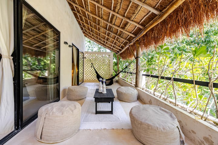 Une terrasse extérieure avec un toit de chaume, des poufs beiges, une table basse noire et un hamac, dans un cadre verdoyant baigné de lumière naturelle.