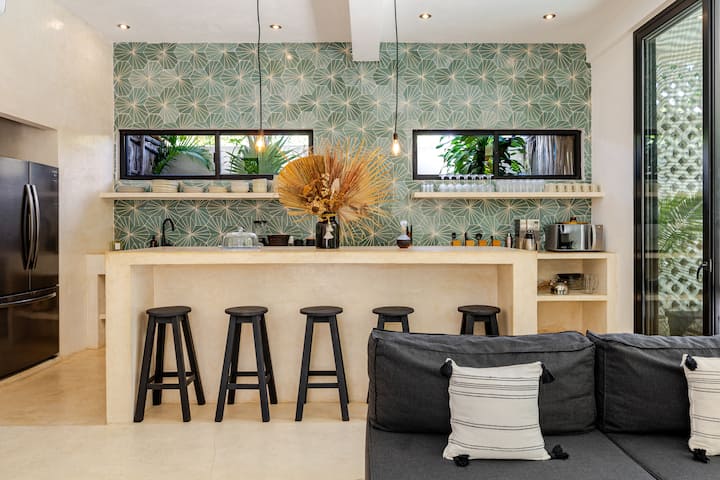 Une cuisine moderne au design tropical avec un plan de travail beige, des tabourets de bar noirs, un mur aux motifs verts et des plantes décoratives visibles à travers des fenêtres étroites.