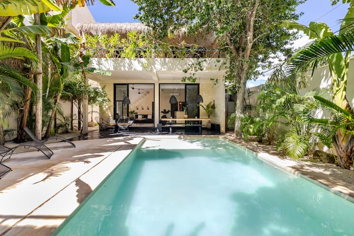 Un logement tropical moderne avec une piscine privée entourée d'une végétation luxuriante, un salon d'extérieur et de grandes portes-fenêtres menant à un espace de vie lumineux.