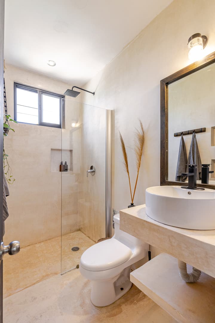 Salle de bain lumineuse aux murs beiges avec cabine de douche vitrée, ciel de douche noir, lavabo en céramique blanche et décoration minimaliste avec des herbes de la pampa et des serviettes suspendues à un porte-serviettes en bois.