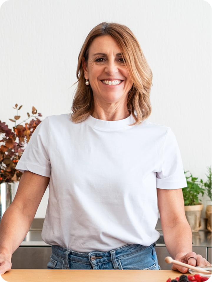 Deborah Compagnoni stands in a chefs kitchen wearing a white shirt and jeans and smiles into the camera over composed plates of food