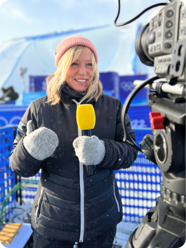 Reporter Jenny Jones holds a larger yellow microphone and speaks to camera reporting from a ski slope